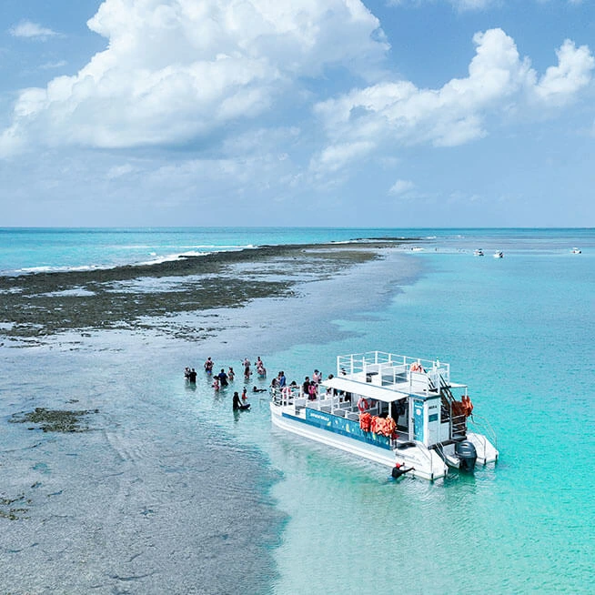 Catamarã ancorado nas piscinas naturais de Japaratinga, com águas cristalinas e turistas explorando o local em um dia ensolarado.