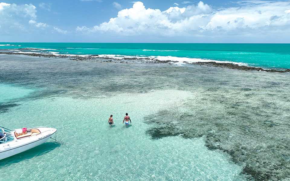 Casal caminha dentro da água cristalina nas piscinas naturais de Japaratinga, desfrutando um cenário paradisíaco, ao lado dos recifes de corais e da lancha de passeio.