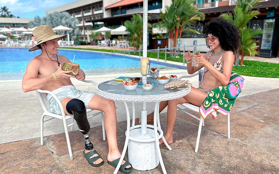 Casal vestido com roupas de banho desfruta de drinks e petiscos em mesa branca ao lado da piscina do Verano.