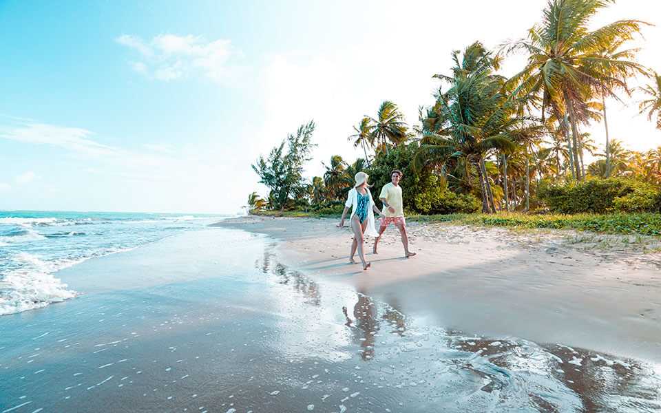 Casal caminha na areia, entre o mar e a vegetação, na Praia do Toque, em Alagoas.